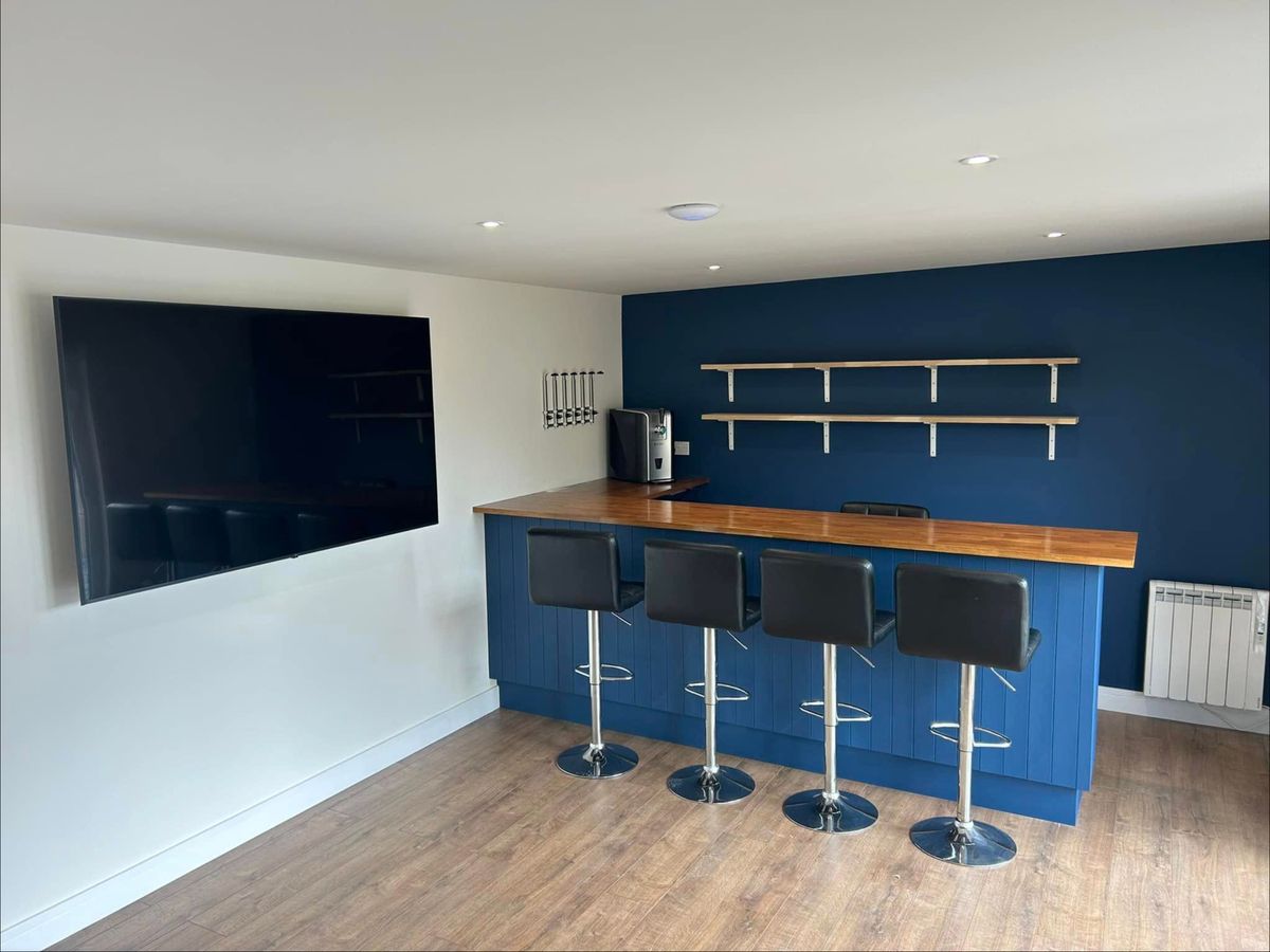 Blue panelled bar with a timber worktop, four bar stools, open shelves and a wall mounted TV
