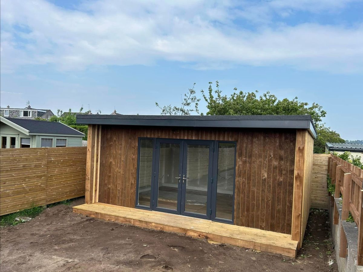 Garden room clad in vertical timber boards with grey doors and a deck, on bare soil