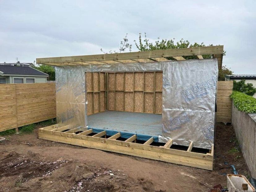 Timber frame of a garden room on its base, partly wrapped in membrane