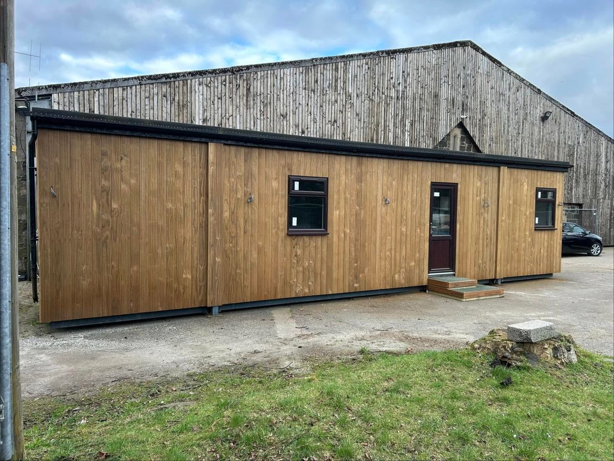 Long portacabin clad in vertical thermowood boards, with dark framed windows and a door with steps